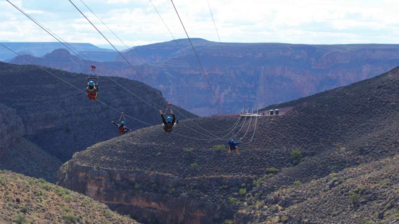 Four people are seen zip-lining across a deep, V-shaped canyon gorge with steep, rocky walls, against a backdrop of distant, hazy blue mountains.