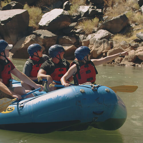 A group in helmets and life vests on a blue raft, with one person pointing ahead.