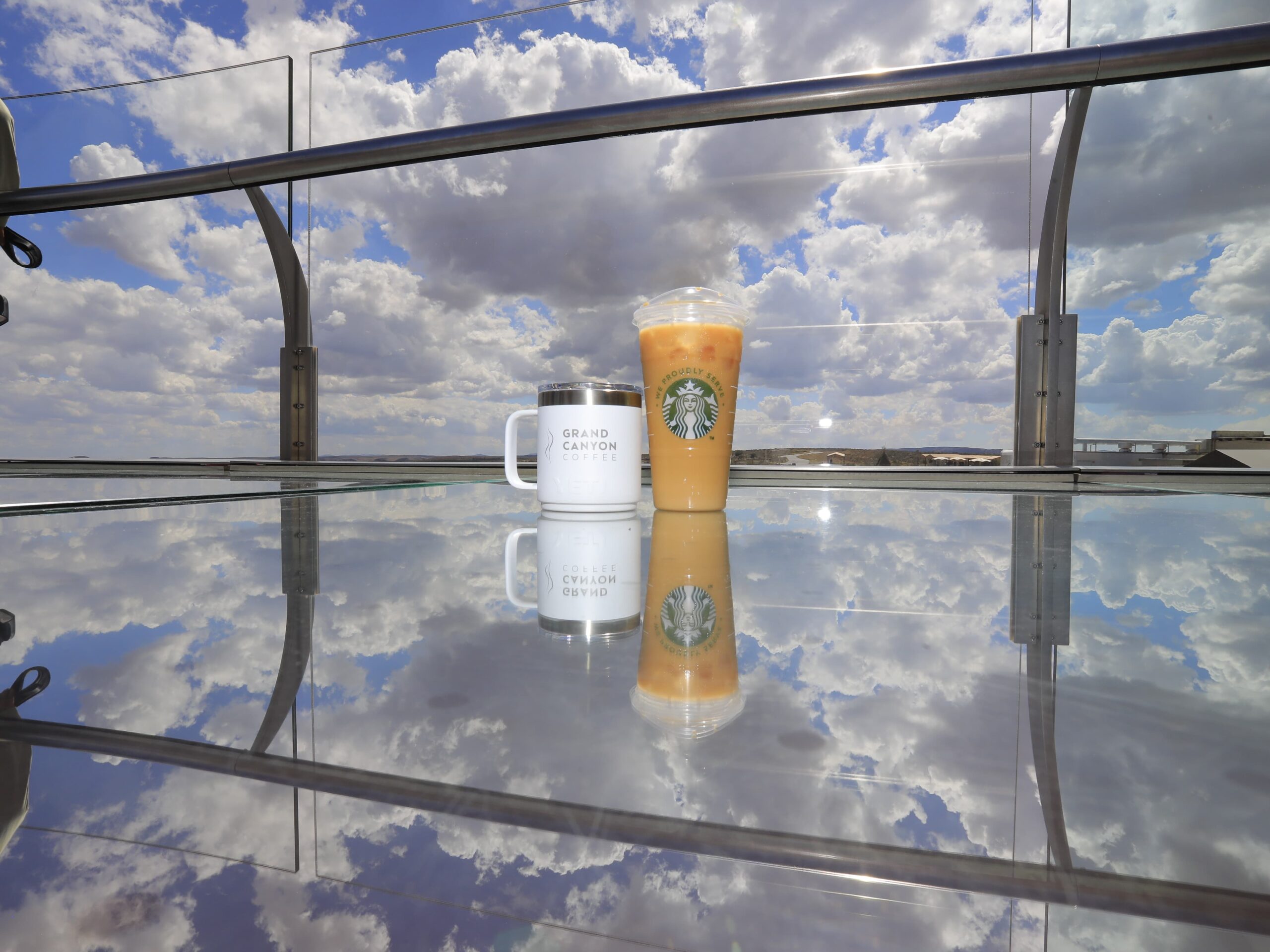 A Grand Canyon Coffee mug and a Starbucks iced coffee sit on the glass floor of the Grand Canyon Skywalk, with the cloudy blue sky perfectly reflected on the surface