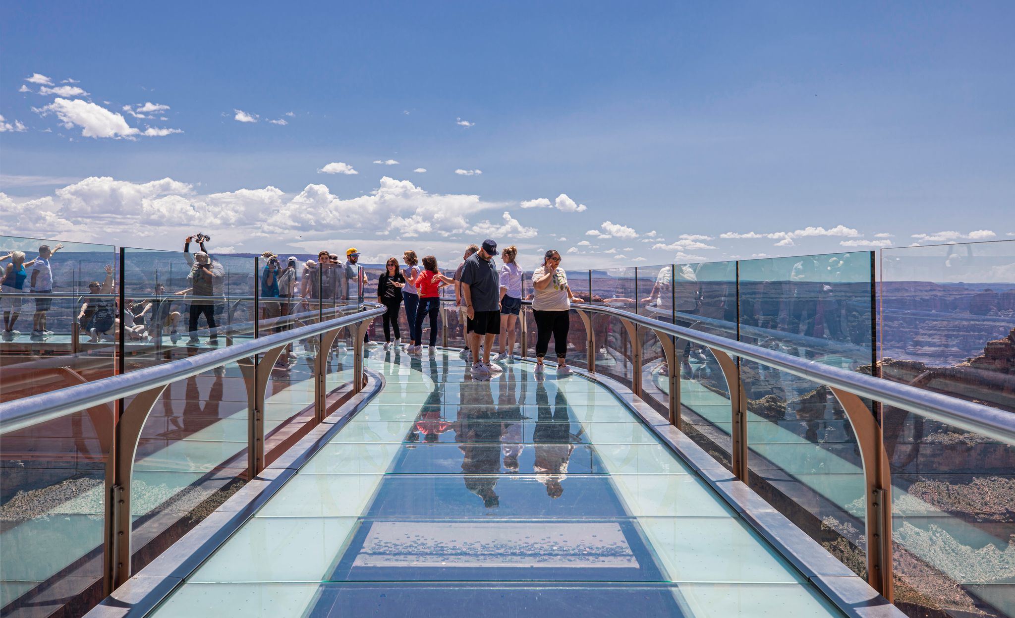 Phoenix to Grand Canyon Road-Trip Guide A group of tourists is walking on the U-shaped, glass-floored Skywalk over the edge of the Grand Canyon, which offers panoramic views of the distant canyon landscape under a bright blue, cloudy sky.