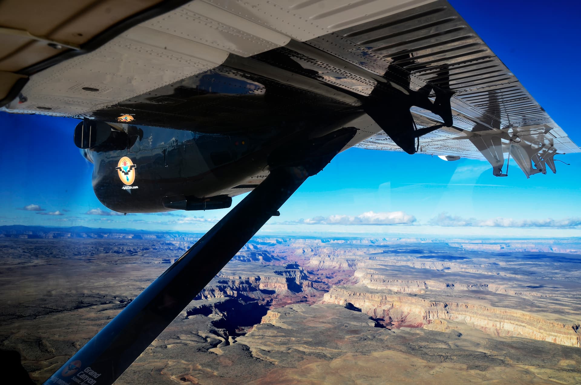 Ventes de groupe An aerial view of the Grand Canyon taken from the window of a sightseeing airplane. The plane's wing is visible in the foreground, with the vast, layered canyon landscape unfolding below under a deep blue sky.