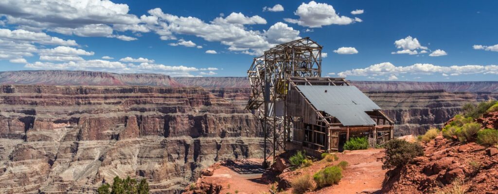 An old mining structure sits on the edge of the Grand Canyon under a cloudy blue sky.