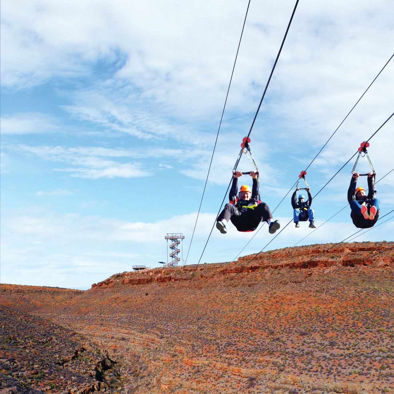 Three individuals, wearing helmets and harnesses, are suspended in the air while riding parallel ziplines. They appear to be in mid-flight over a canyon or gorge.