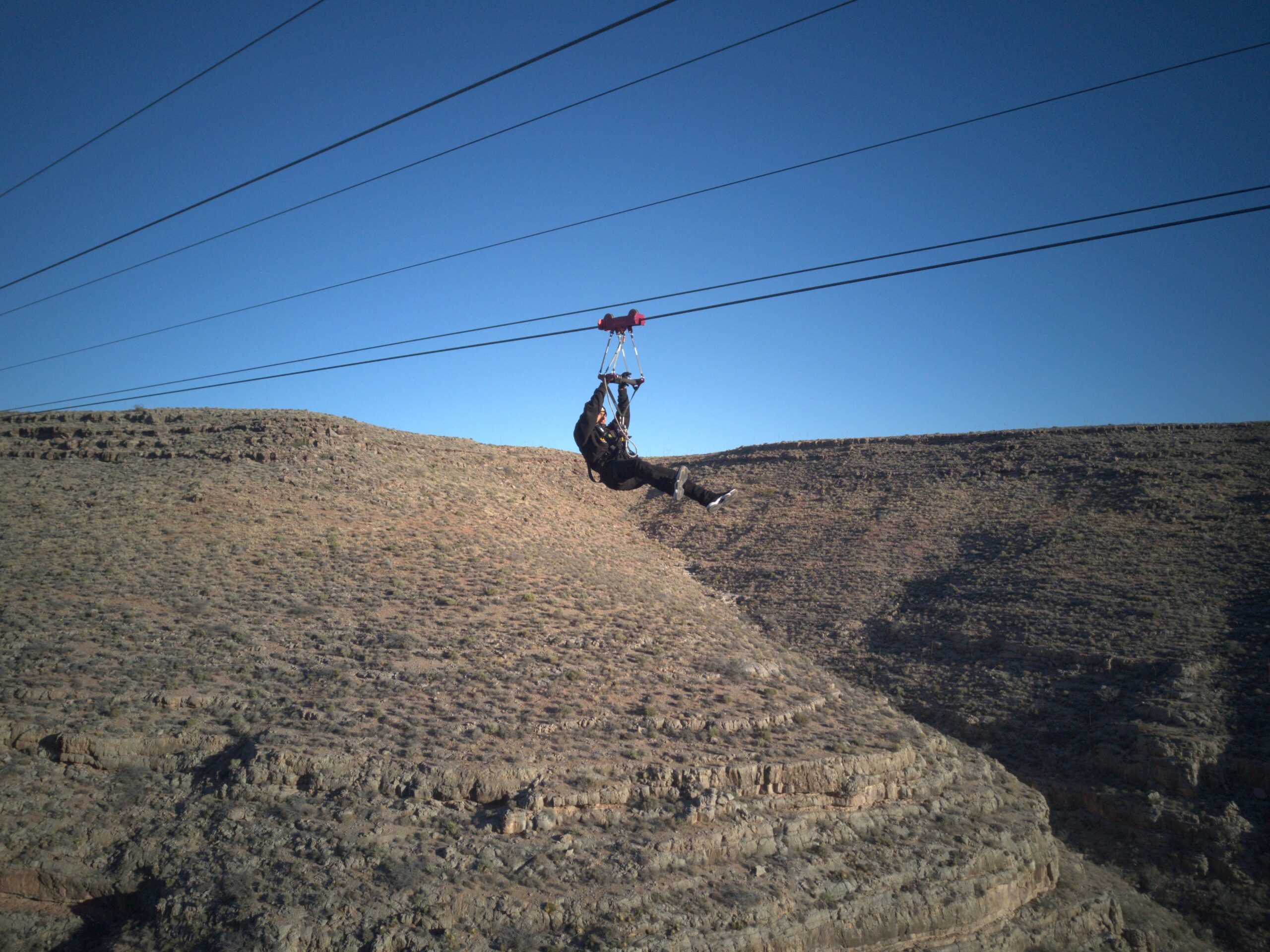A person in dark clothing and a helmet is seen mid-flight on a zipline, suspended over a rugged, rocky canyon beneath a clear blue sky.