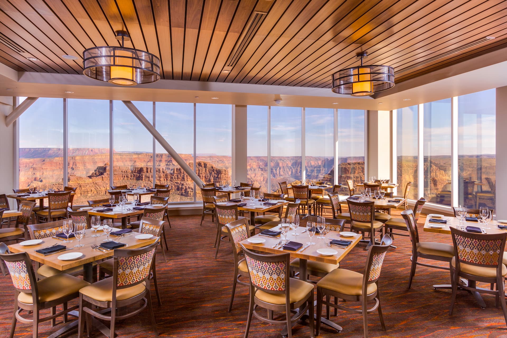 Ventes de groupe The interior of a restaurant with floor-to-ceiling windows that offer a stunning panoramic view of the Grand Canyon. The dining room is elegantly set with tables and chairs, awaiting guests.