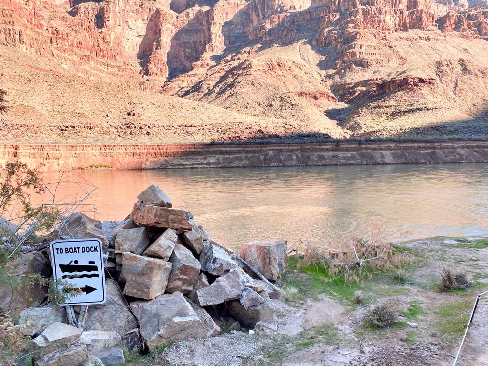 A sign pointing to a boat dock sits on a pile of rocks beside a river, with the Grand Canyon walls in the background.