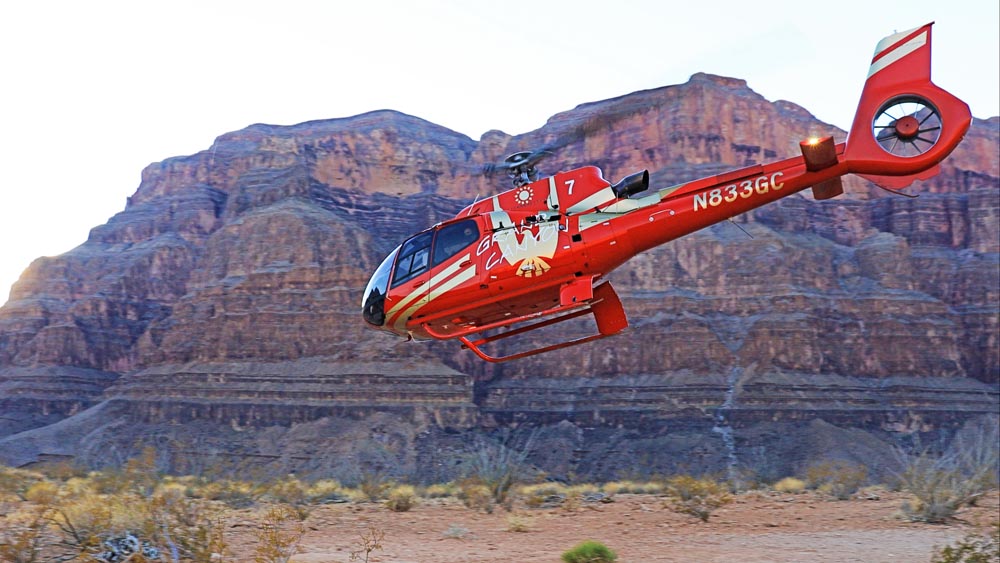 A red helicopter is shown mid-air, with the Grand Canyon in the background.