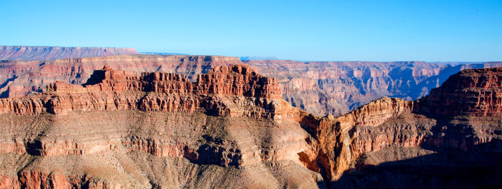 A panoramic view shows the layered, reddish rock formations of the Grand Canyon under a clear blue sky.