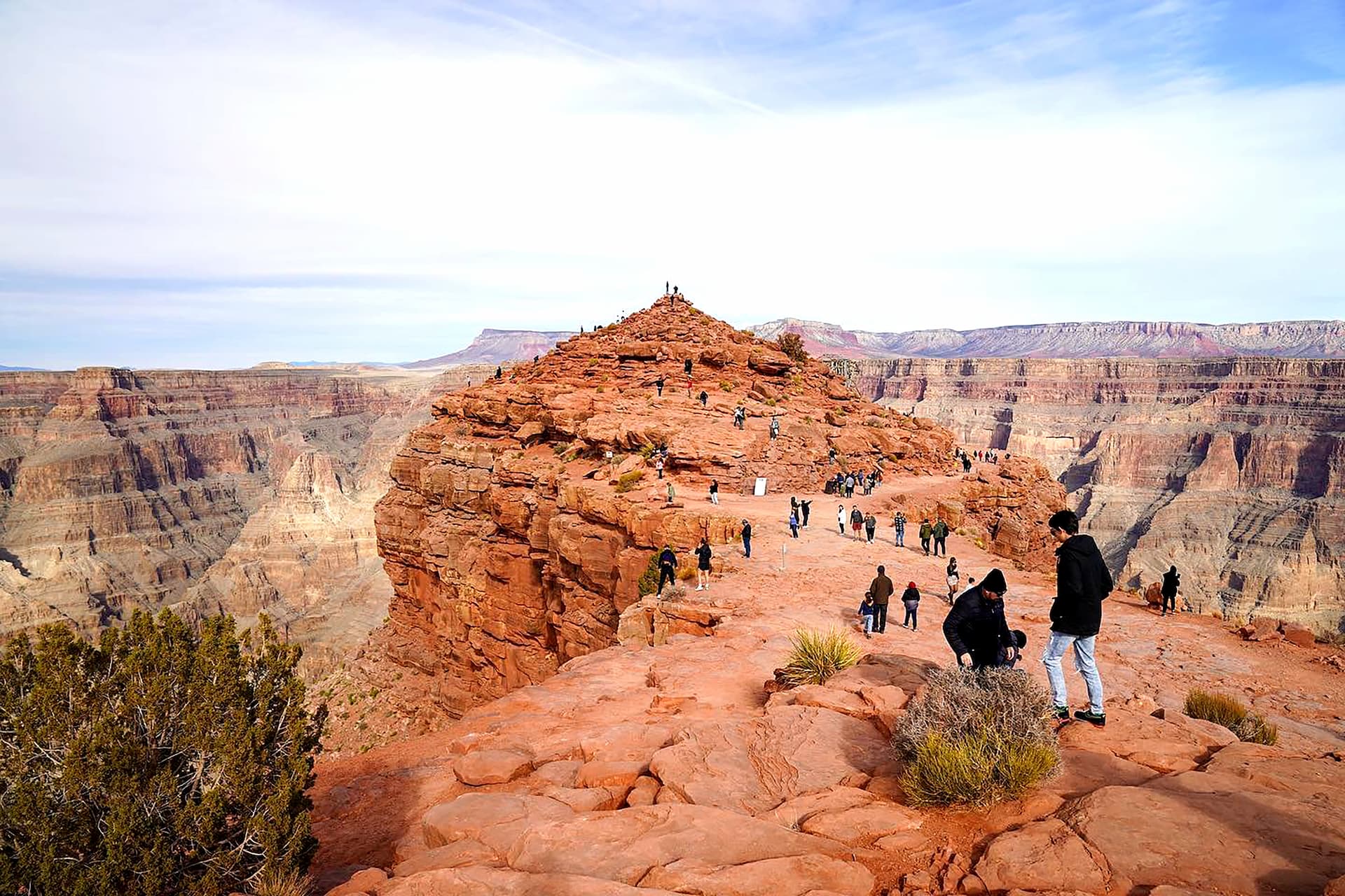Ventes de groupe Tourists explore Guano Point, a large red rock viewpoint that extends into the Grand Canyon. The vast, layered canyon walls stretch into the distance under a bright sky.