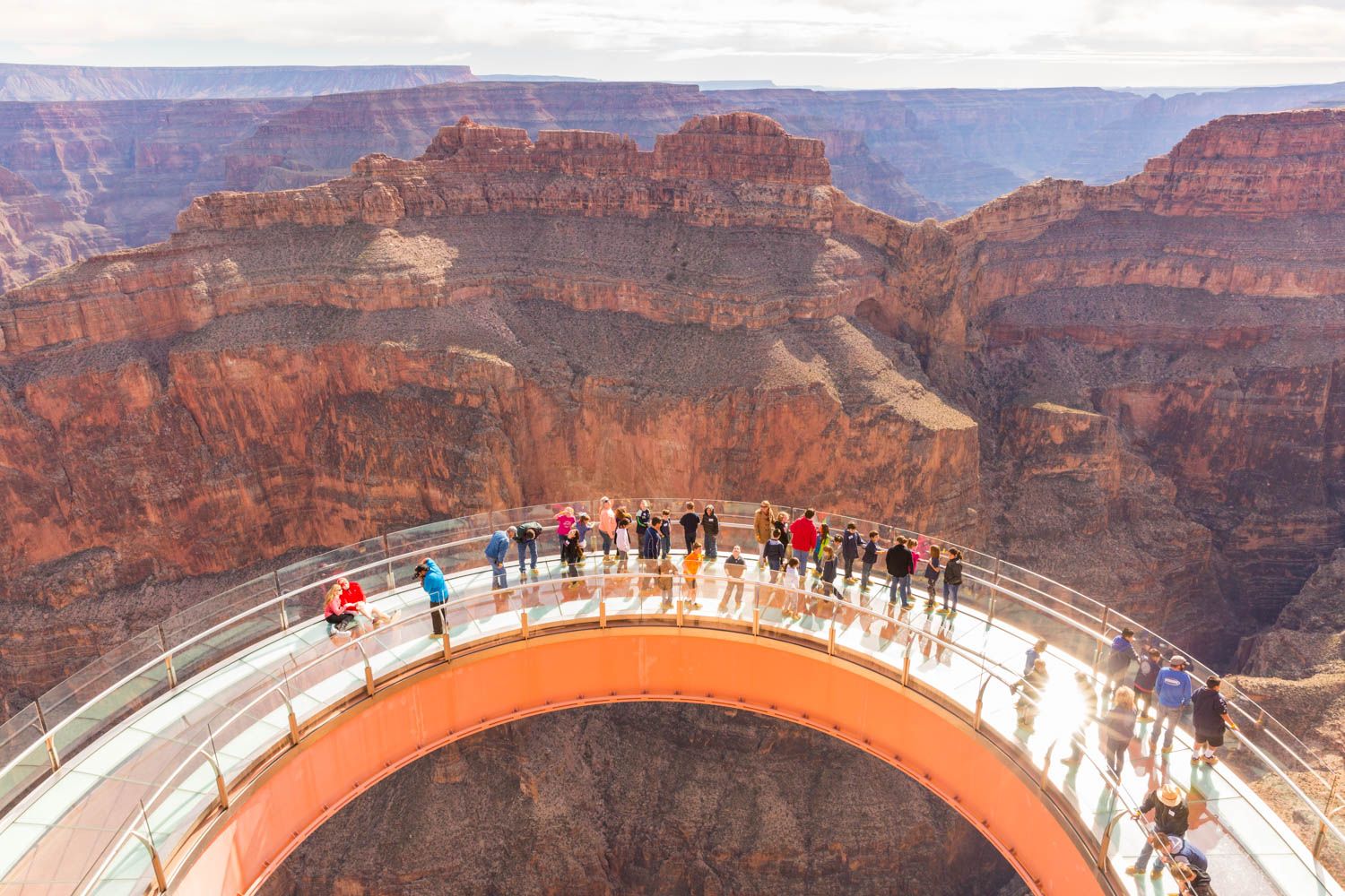 Ventes de groupe A high-angle view of tourists standing on the Grand Canyon Skywalk, a large, horseshoe-shaped glass bridge extending over the vast, layered red rocks of the canyon.