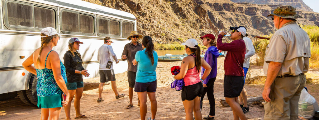 A group of people are gathered on a dirt bank next to a white bus on a tour of the Grand Canyon