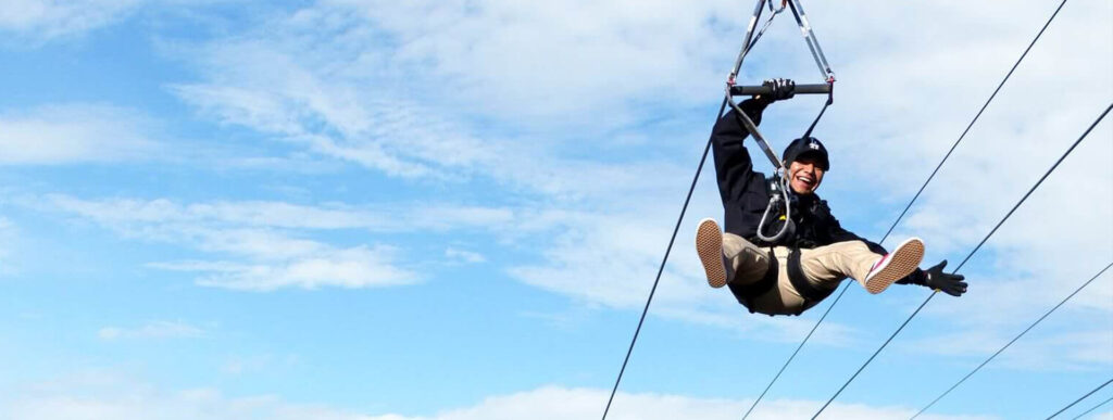 Choses à faire A person smiles while zip-lining against a blue, cloudy sky.