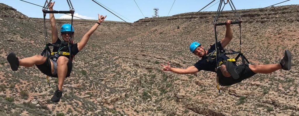 Two people on zip lines smile and pose over a desert canyon.