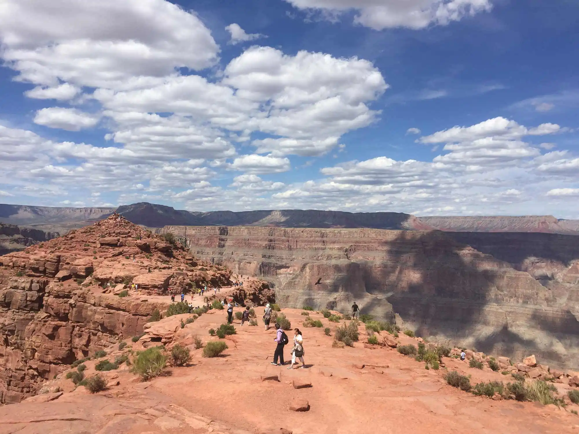 Tourists walk on a rocky path overlooking a vast canyon under a cloudy blue sky.